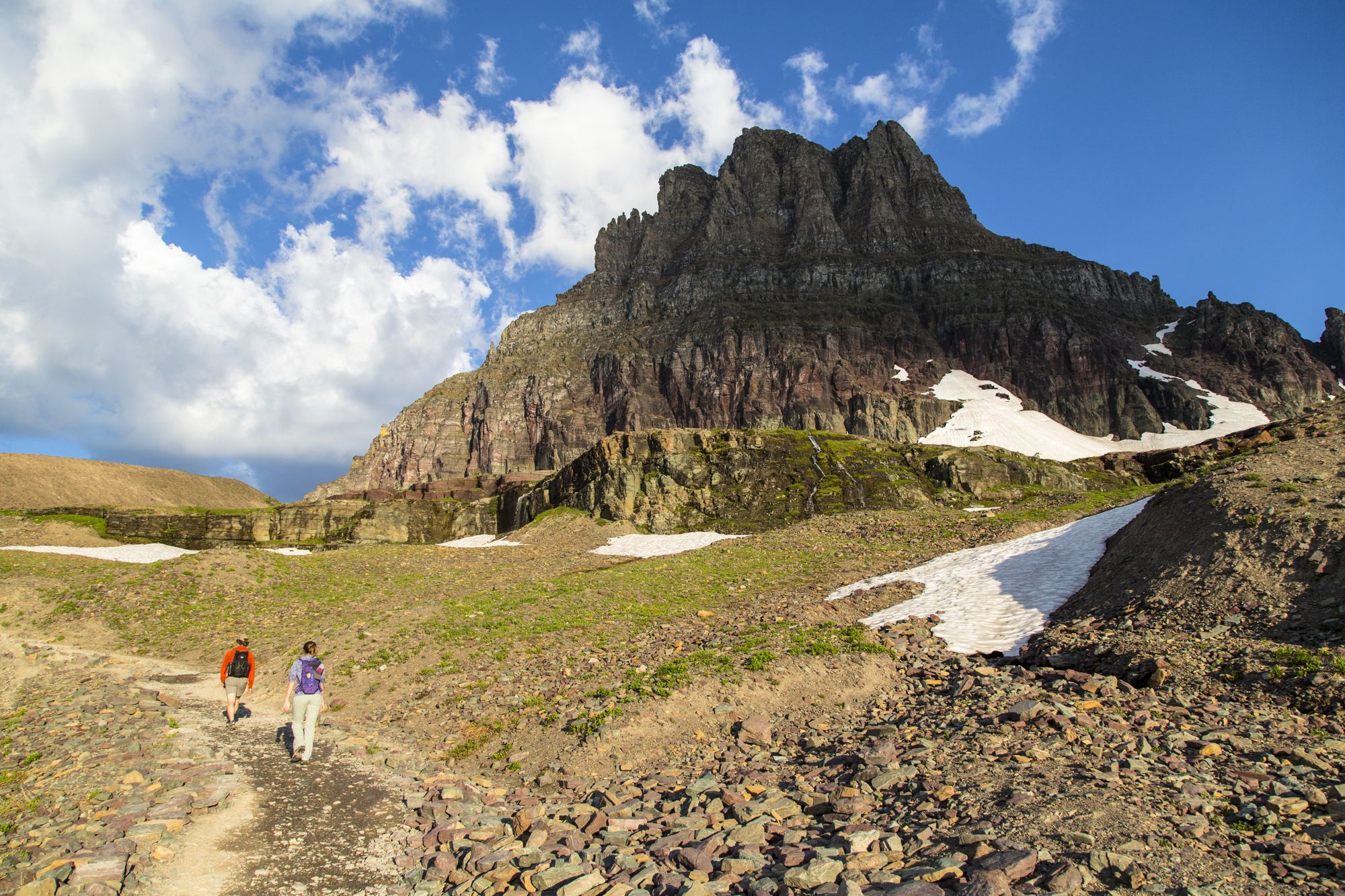 Two hikers on a rocky trail below a towering mountain peak